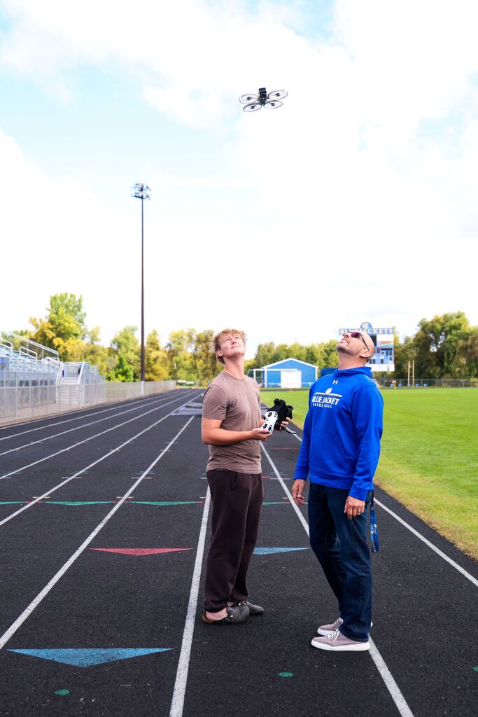 A student and teacher wearing a Bluejacket Basketball sweatshirt look up at a flying drone on a running track.