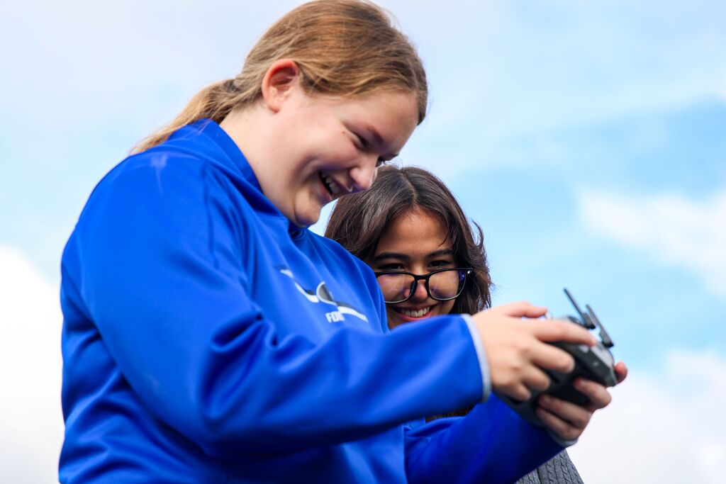 Two female students smile as they hold and look down at a drone controller outdoors.