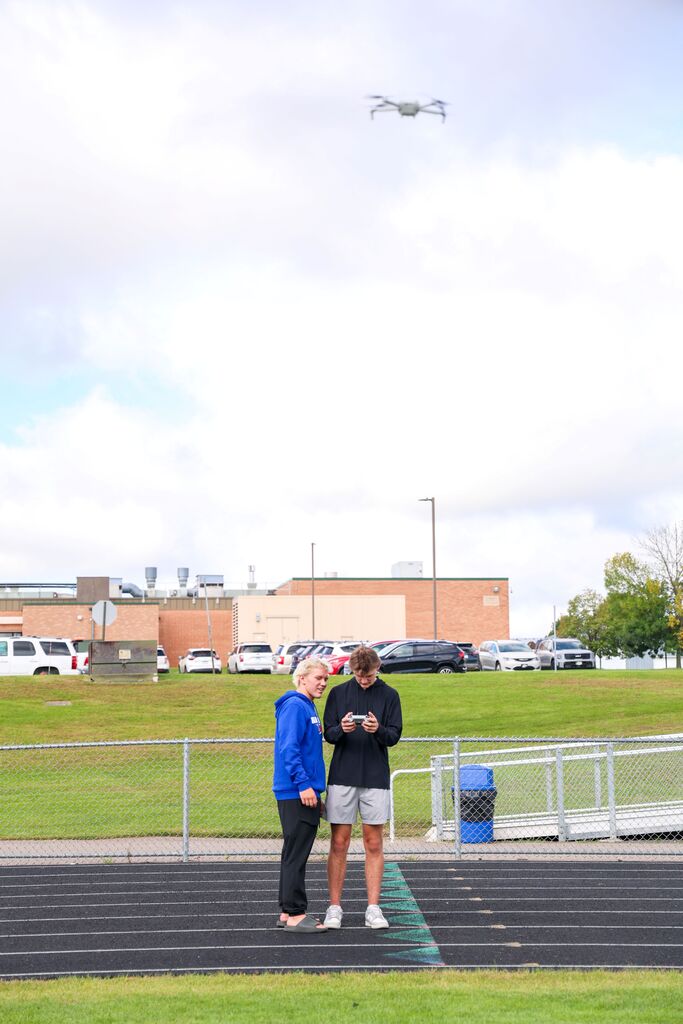 Two students stand on a track and operate a drone controller while looking up at a flying drone.