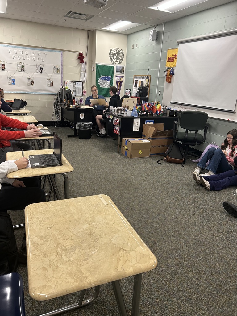 student sit on floor against a wall working together, which a separate student meets with the teacher to discuss any help that may be needed on coursework.