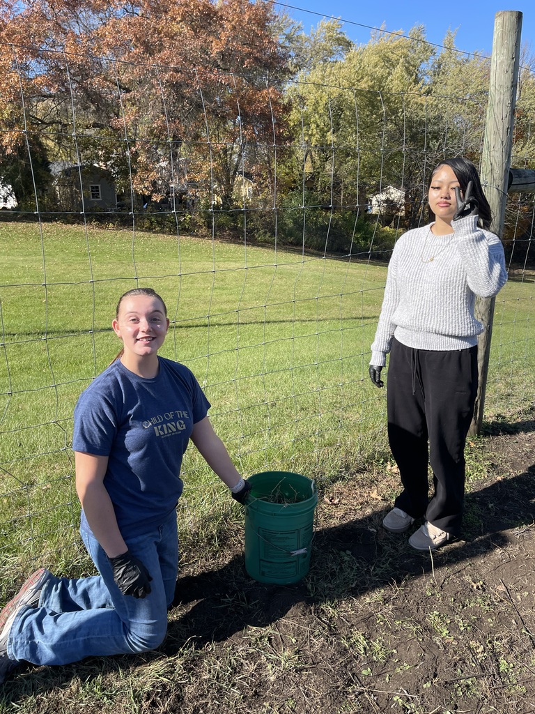 Two students participate in an garden activity next to a green bucket along a wire fence.