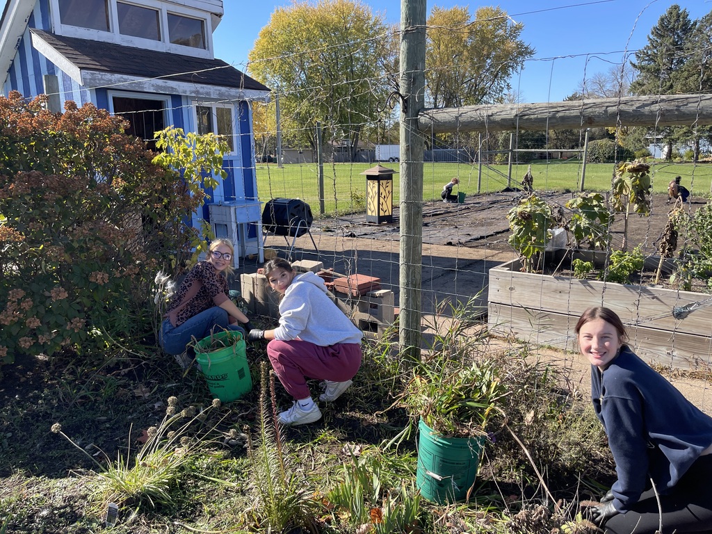 Three students crouch outdoors in a garden area, putting weeds into green buckets next to a small blue shed.