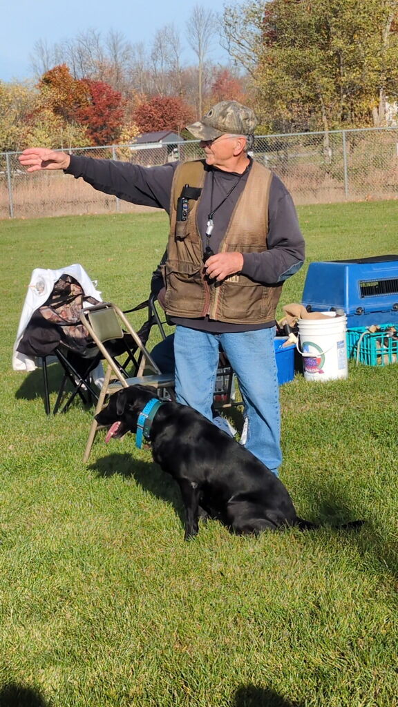 An instructor in a camouflage hat gestures while a black dog sits beside him on a grassy field.
