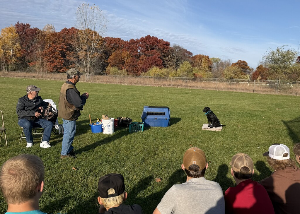 Students sit on a field, watching an instructor present a dog training demonstration with a black dog sitting on a mat.