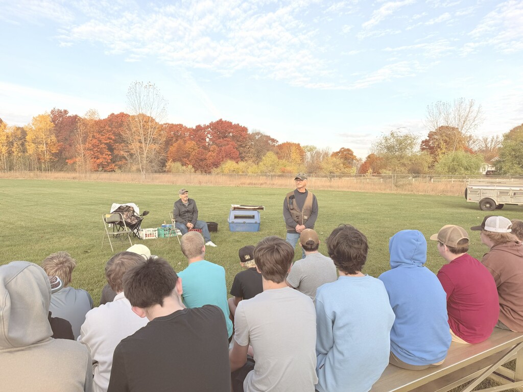 Students sit on bleachers outdoors while a speaker presents a dog training demonstration on a field.