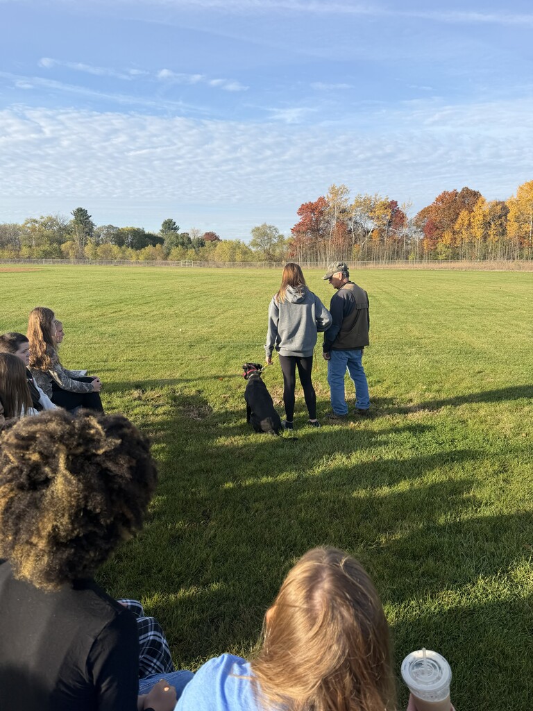 Students sit on an outdoor field watching a demonstration with a black dog, trainer, and handler.