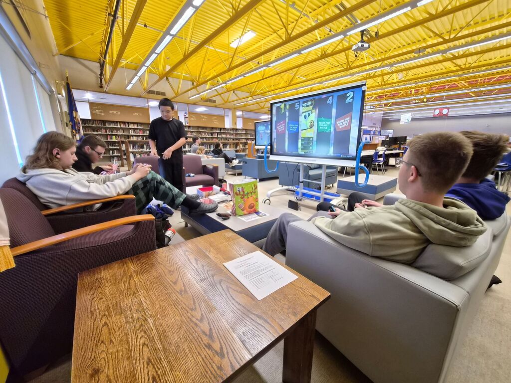 Students sit on armchairs in a library lounge area while playing a video game displayed on a large screen monitor.