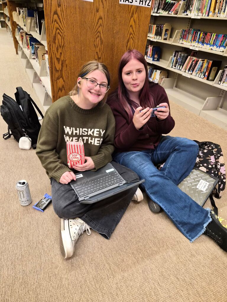 Two students sit on a library floor, working on a laptop and using a phone with snacks nearby.