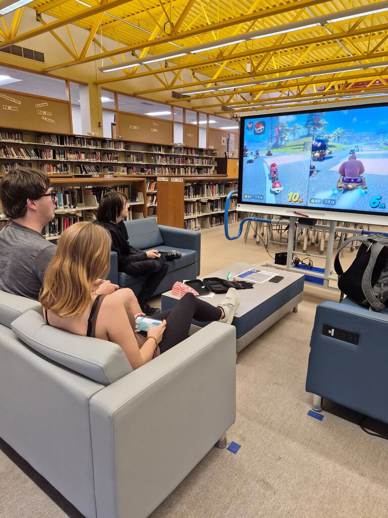Students sit in a library lounge area playing a video game on a large screen display.