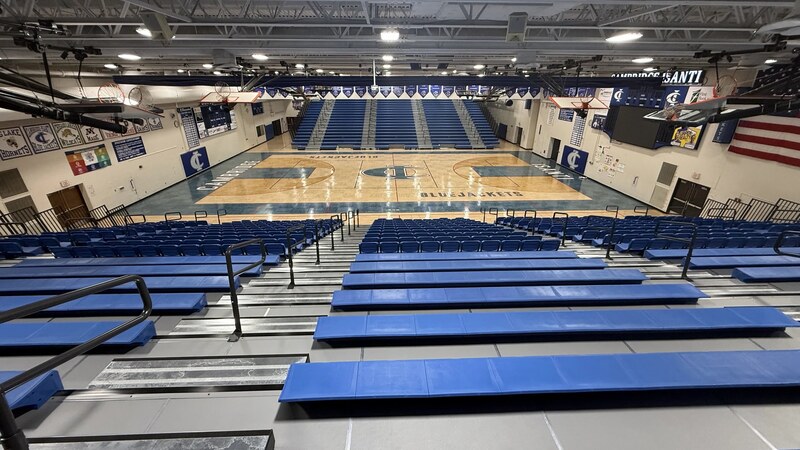 Empty blue bleachers face an empty basketball court in a school gymnasium.
