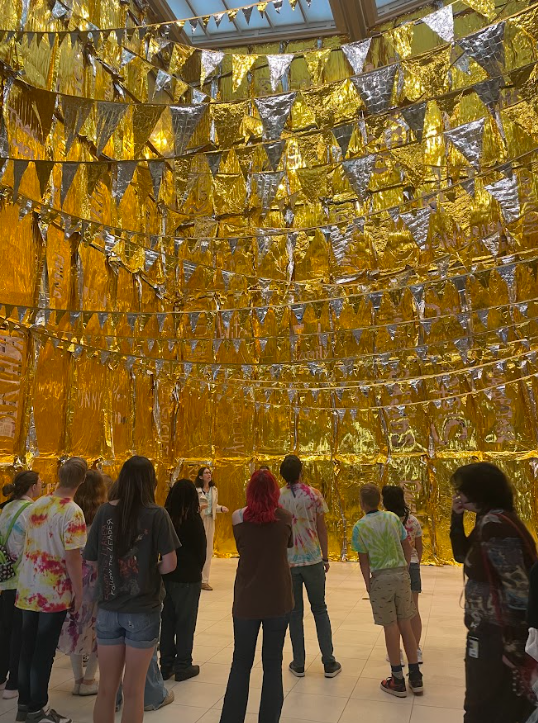 A group of students seen from behind gazing up at gold flags that line the ceiling of a room.