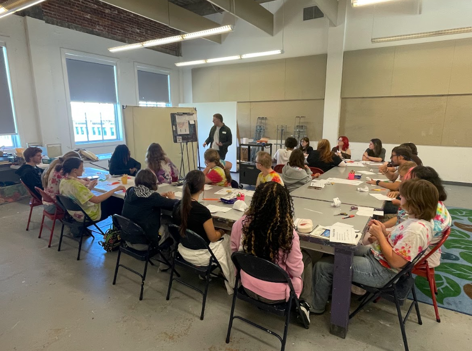 A group of students sitting around a table with art supplies in front of them and being directed by an artist .