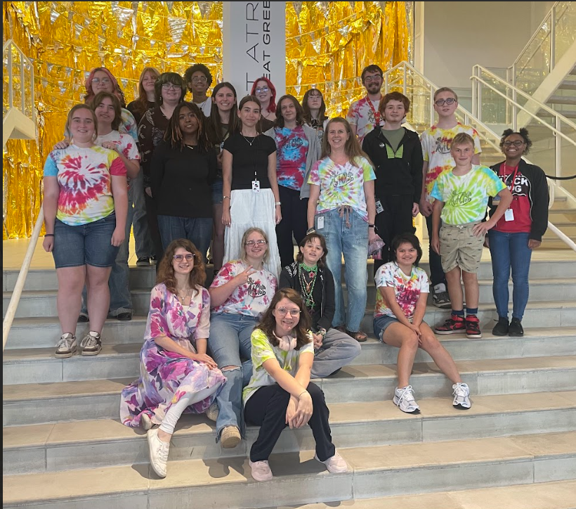 Art Club students posing on a staircase in front of a Gold backdrop. Some students are standing while some are sitting on the stairs.
