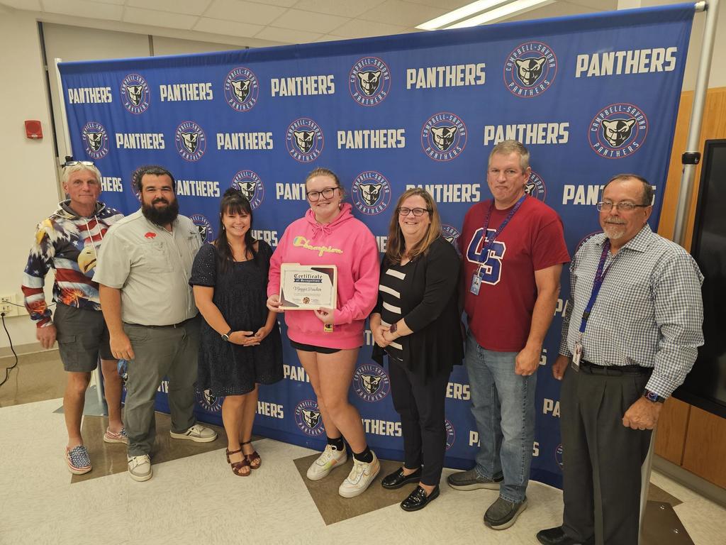 Board of Education posing in front of banner with student in pink holding certificate of recognition.,