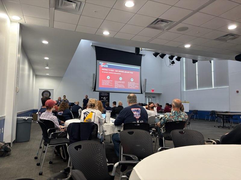 Picture of students and adults looking at screen with red background and information about drones.