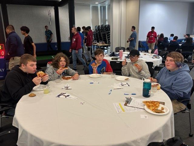 Group photo of students eating pizza at table.