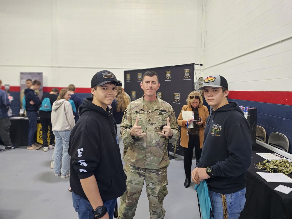 2 students with baseball caps posing for picture with army recruiter