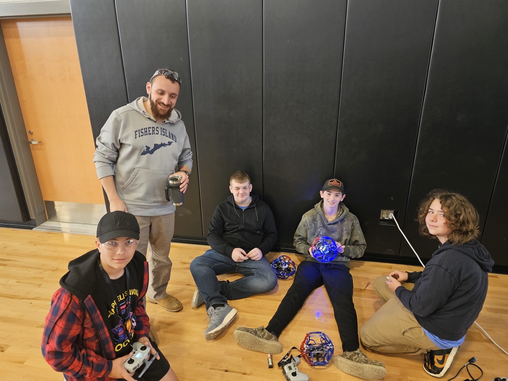 4 students and coach posing for picture while sitting on the gym floor working on drones.