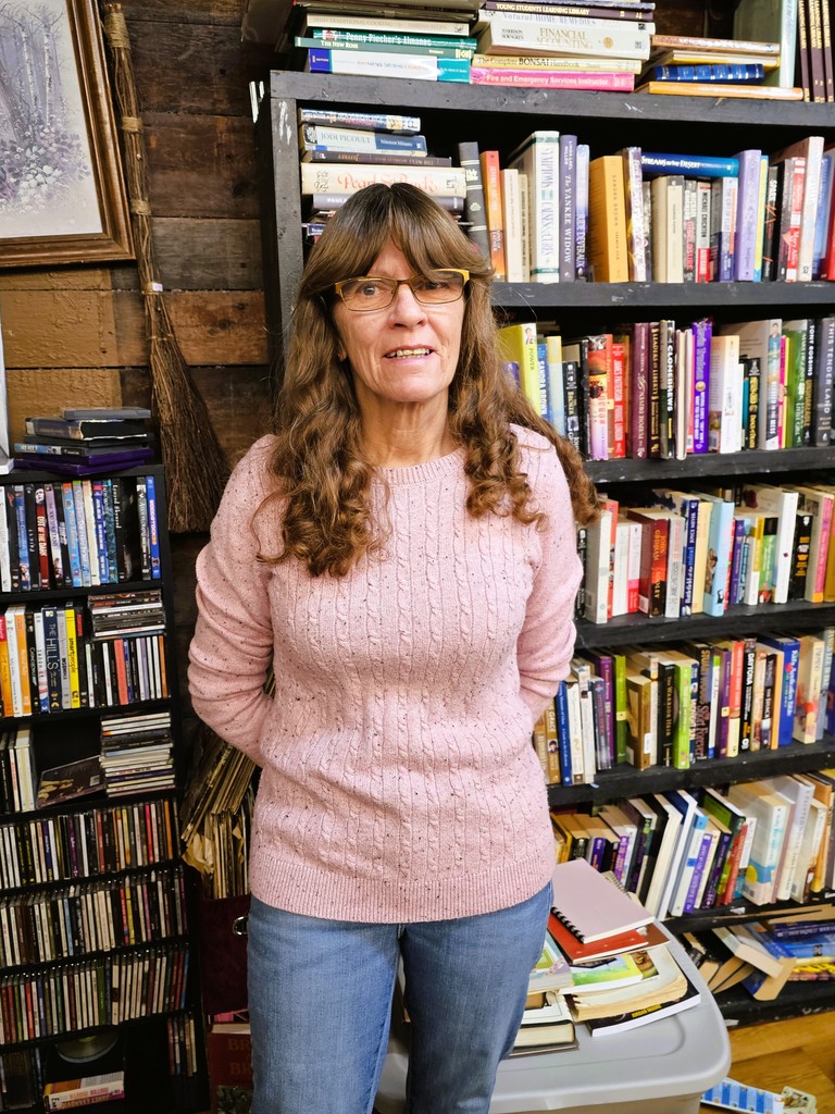 Delinda Newman wearing pink sweater, jeans standing in front of a bookshelf