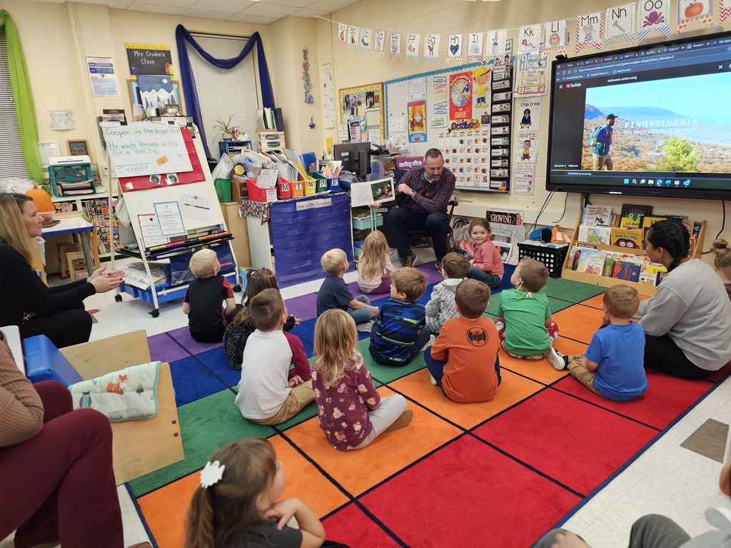 PreK students sitting on rug while adult in flannel shirt reads a story to them