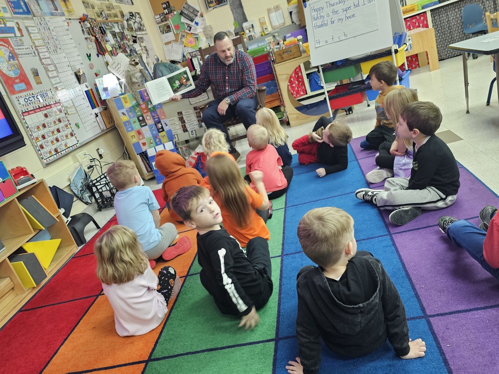 PreK students sitting on rug while adult in flannel shirt reads a story to them