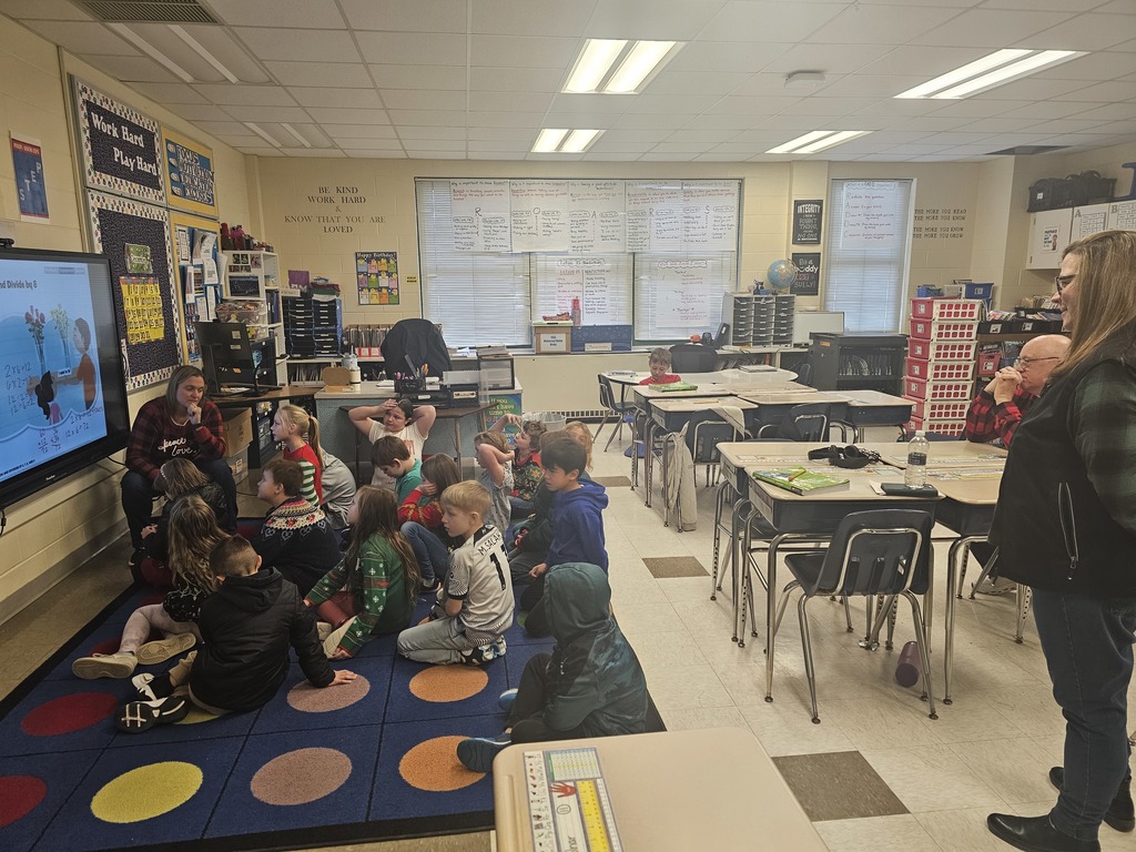 Board of education visting classroom, students sitting on rug with teacher