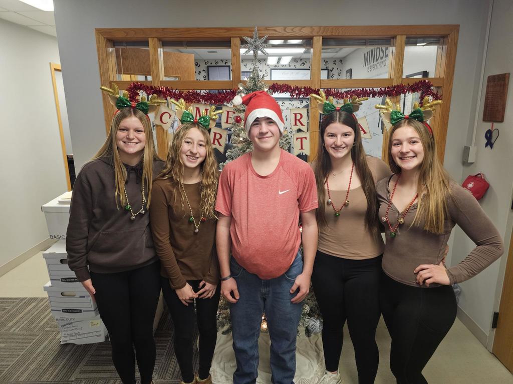 Five students posing in front of a christmas tree 4 dressed as reindeer one as santa