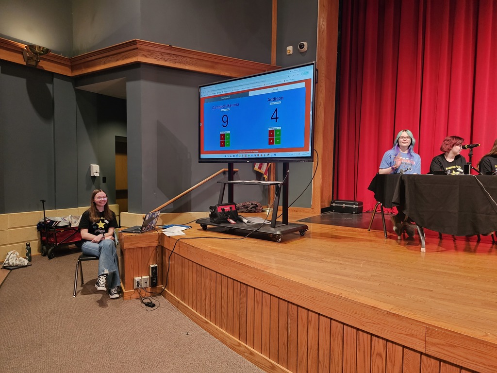 Student sitting side stage at the computer with the Academic all stars score on a smarboard.
