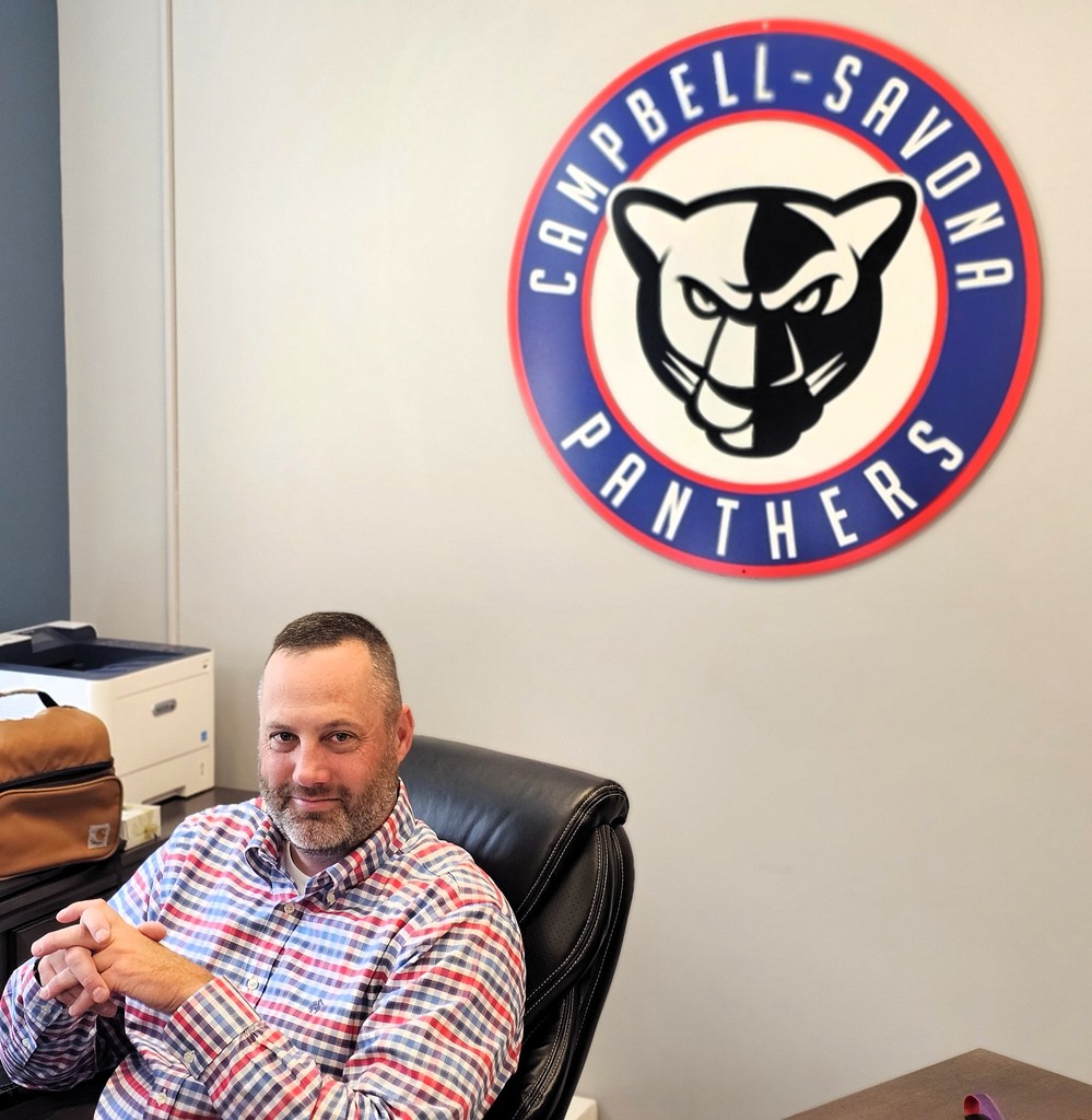 Supt. pic sitting in chair in office with school emblem in background