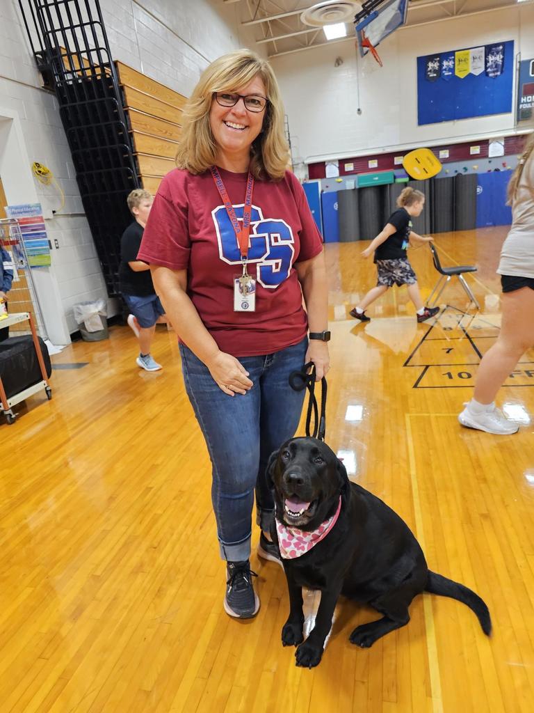 Teacher in maroon school shirt with black lab on leash sitting next to her.
