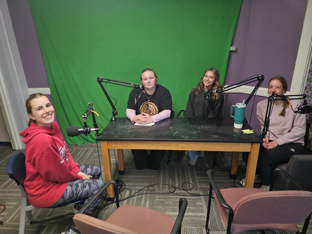 4 students sitting in front of microphones and green screen