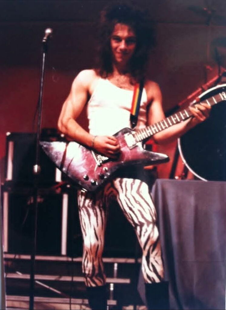 Man with purple gibson explorer, teased black hair in white tank top and spandex leggings.