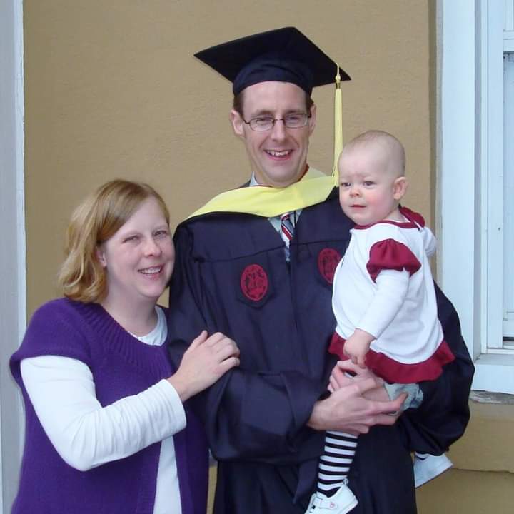 Man in graduatiion mortar board holding baby with wife by his side