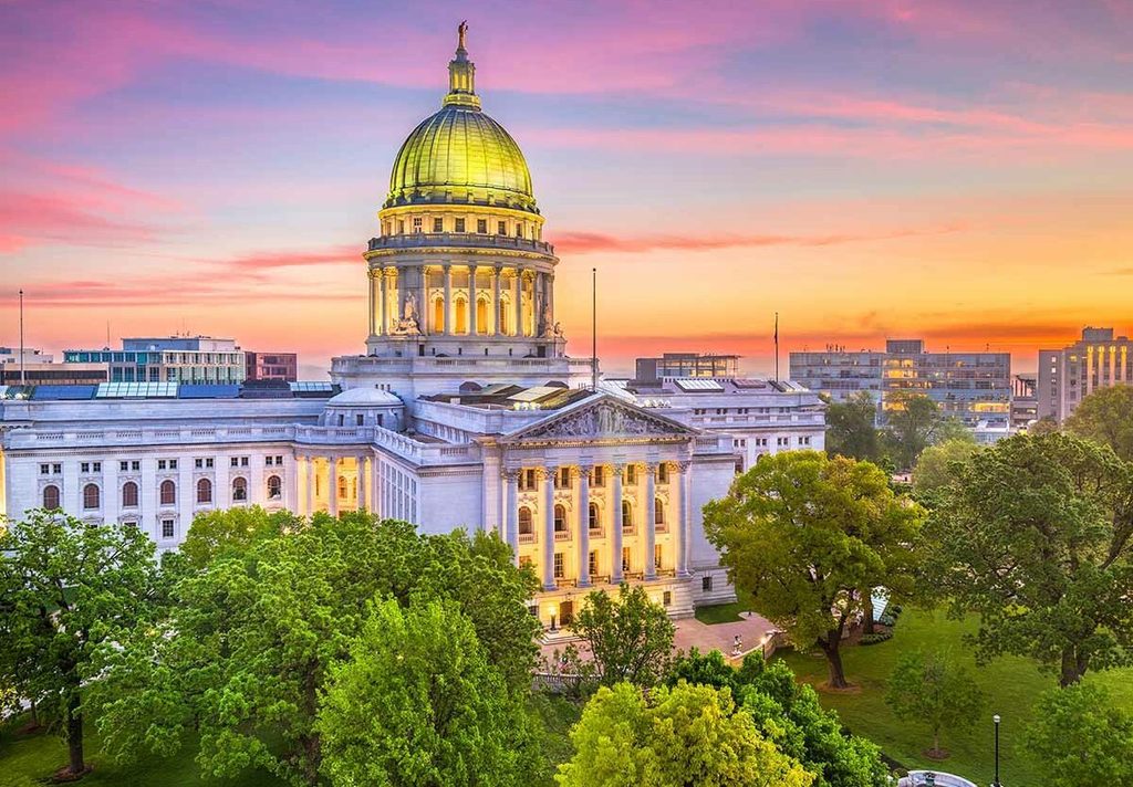 Picture of the Capital in Madison Wisconsin with a beautiful sunset in the background with green trees in the forefront.