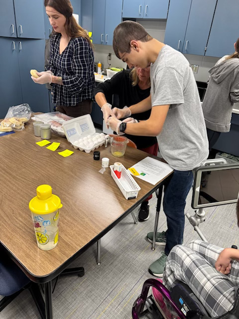 Picture of a student cracking eggs to make a breakfast sandwich while a teacher is cutting the english muffins.