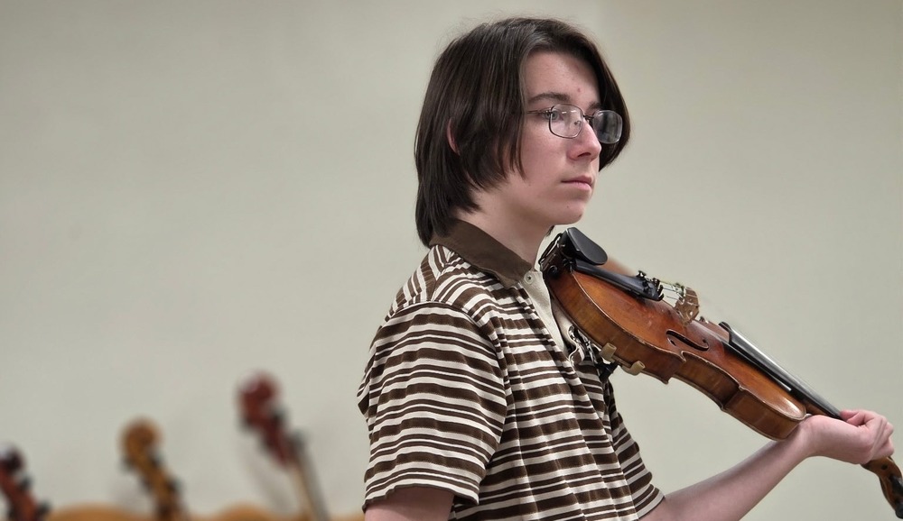 A young musician with glasses wearing a striped shirt plays a violin indoors.
