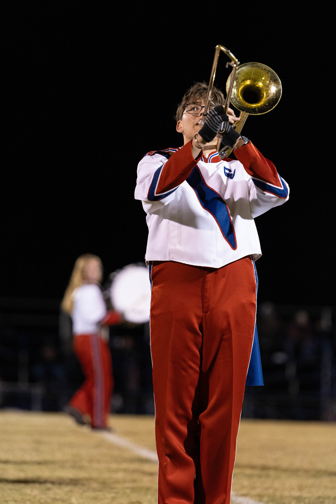 Photo taken looking from the ground up of a student playing an instrument during a football halftime performance. The white uniform with brass instrument is silhouetted against a dark night sky. 