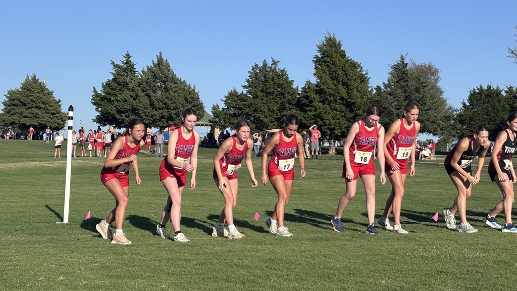 Six Caney girls cross country runners in red uniforms prepare to start a race alongside other competitors on a grassy course.
