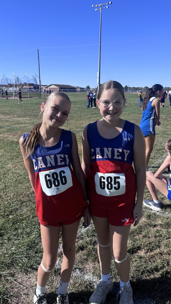 Two Caney cross country runners smile together on a grassy field, wearing blue and red uniforms with race bibs 686 and 685.