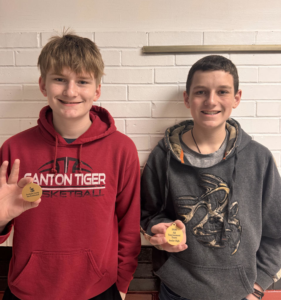 Two middle school boys stand side by side in front of a white brick wall, smiling and holding small gold basketball-shaped medals toward the camera. The boy on the left wears a red “Canton Tiger Basketball” hoodie, and the boy on the right wears a dark hoodie with a deer graphic.