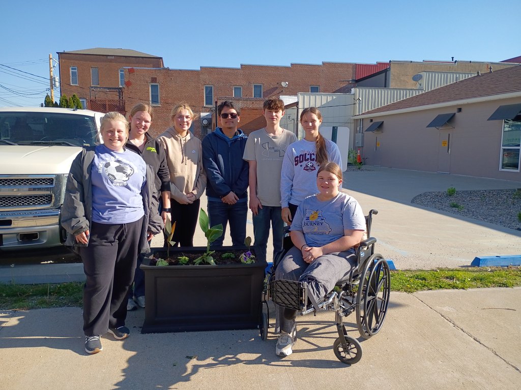 A group of seven high school students pose outdoors on a sunny day behind a large black planter filled with flowers and greenery. One student sits in a wheelchair in front, while the others stand behind her, smiling. A white pickup truck is parked to the left, and brick and metal buildings line the background, suggesting a school or community setting.