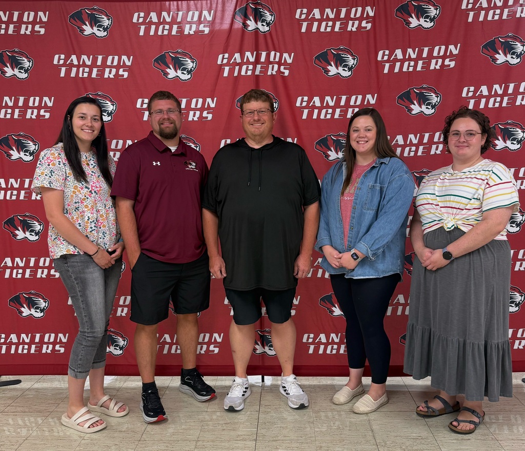 A group of five adults stand side-by-side, smiling and posing for a photo in front of a red Canton Tigers backdrop. The backdrop features repeating white “Canton Tigers” lettering and tiger-head logos in red, white, black, and gray. From left to right: a woman wearing a white floral shirt, gray jeans, and white sandals; a man wearing a maroon Canton Tigers polo, black shorts, and black athletic shoes; a man wearing a black shirt and black shorts with white athletic shoes; a woman wearing a pink shirt, blue denim button-down, black leggings, and light-colored shoes; and a woman wearing a striped white shirt tied at the waist, a long gray skirt, and dark sandals. All five are smiling and standing with their hands relaxed in front of them. The photo is taken indoors on a light-colored tiled floor