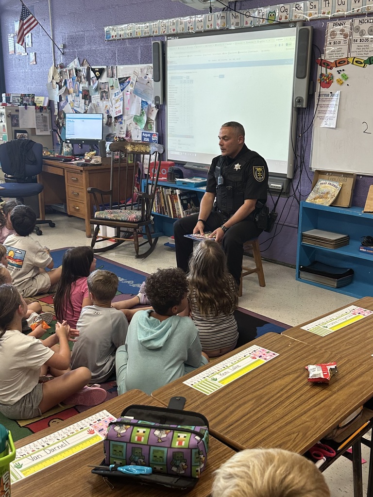 A police officer sits on a stool in an elementary classroom and reads a picture book to several young students seated on a colorful classroom rug.