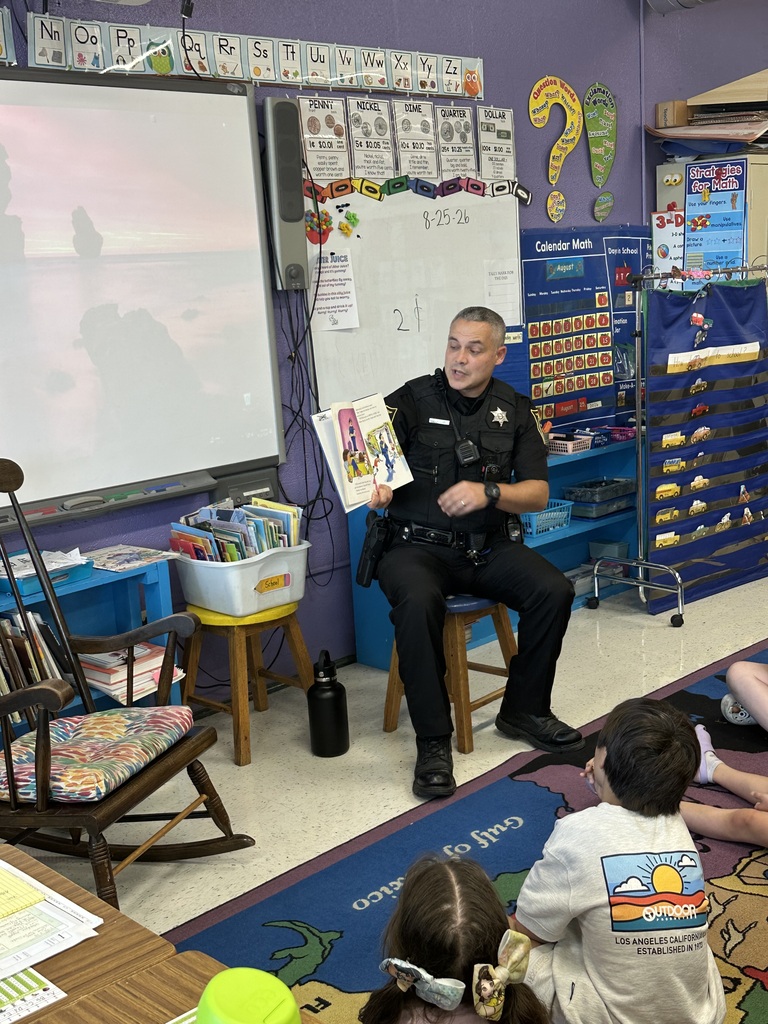 A police officer sits on a stool in an elementary classroom and reads a picture book to several young students seated on a colorful classroom rug.