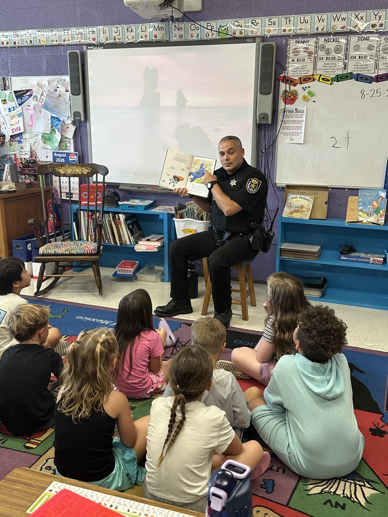 A police officer sits on a stool in an elementary classroom and reads a picture book to several young students seated on a colorful classroom rug.