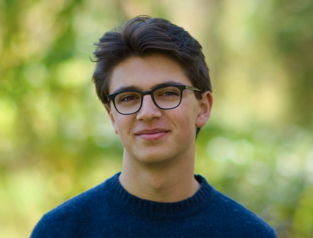Oliver Ames - wearing a blue sweater and glasses, along with a slight smile, Oliver stands in front of a green bokeh background.