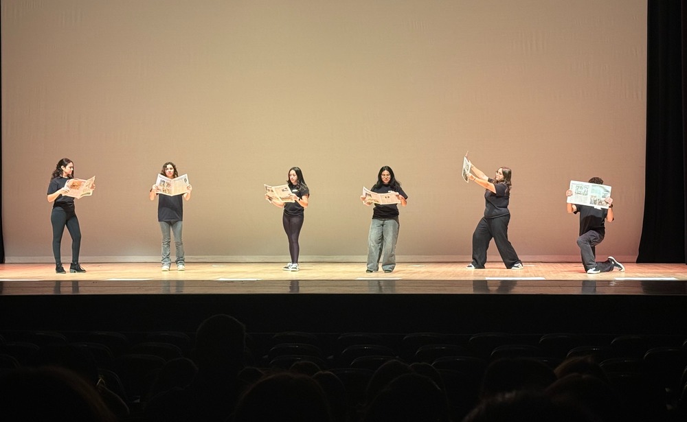 Six performers stand on a stage under warm lighting, each holding a newspaper. Their expressions are focused, conveying an atmosphere of concentration.