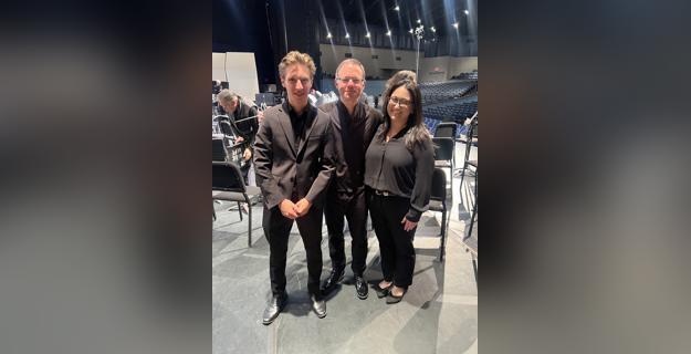 Three individuals dressed in black attire pose together on stage before a performance, with empty seats and stage equipment in the background, capturing a moment of anticipation and collaboration.