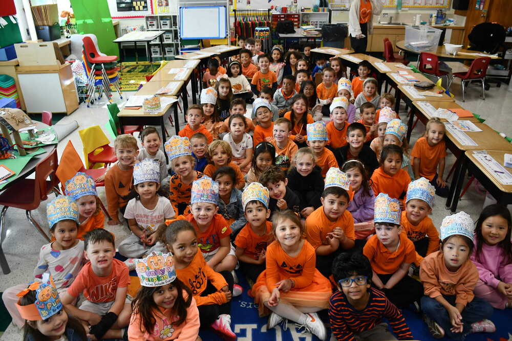 Students sitting on the floor, all wearing orange and paper hats.