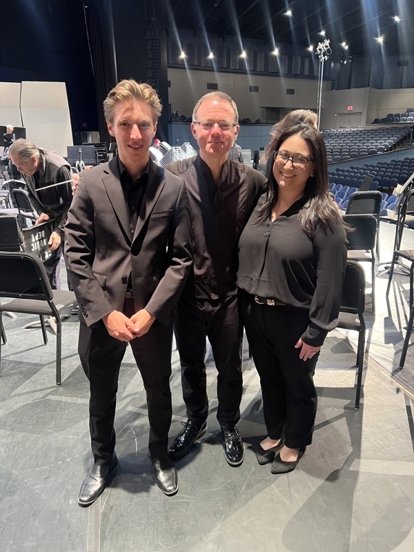 Three individuals dressed in black attire pose together on stage before a performance, with empty seats and stage equipment in the background, capturing a moment of anticipation and collaboration.